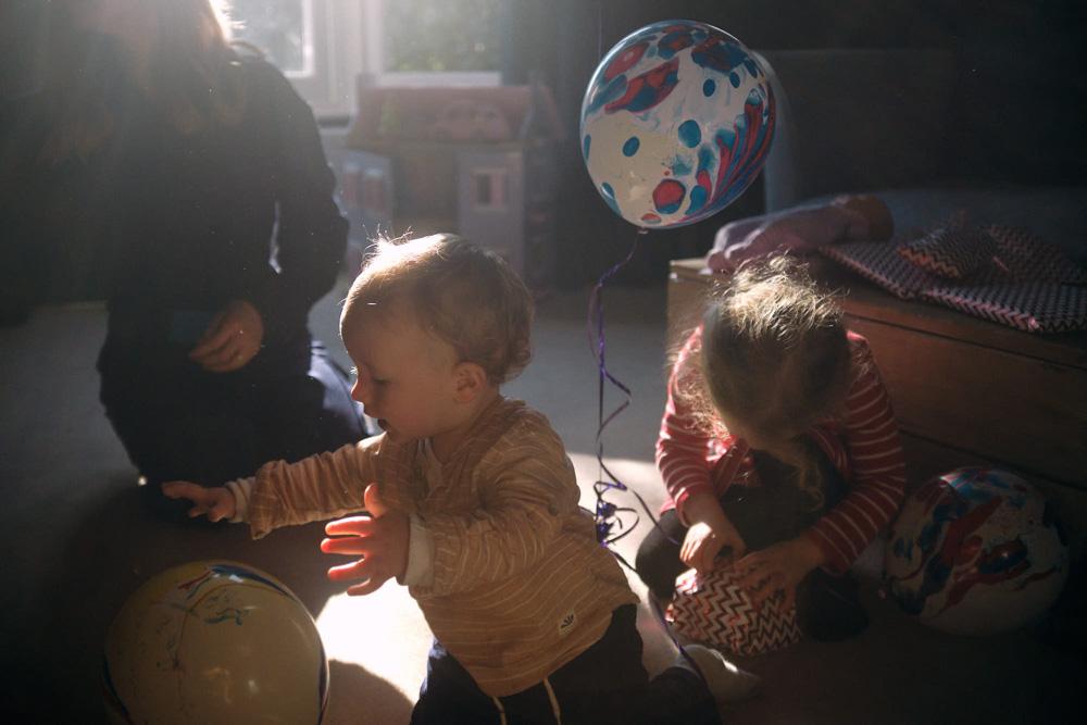 A toddler in a yellow shirt plays with a ball in a sunlit room, while a woman and another child sit nearby. The scene is warm and playful, with a sense of joyful energy.