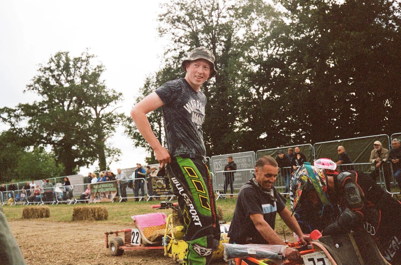 A young man in a bucket hat and camouflage t-shirt stands in front of a crowd of people, with a person in a black t-shirt and orange motorcycle gear to his right. The background.