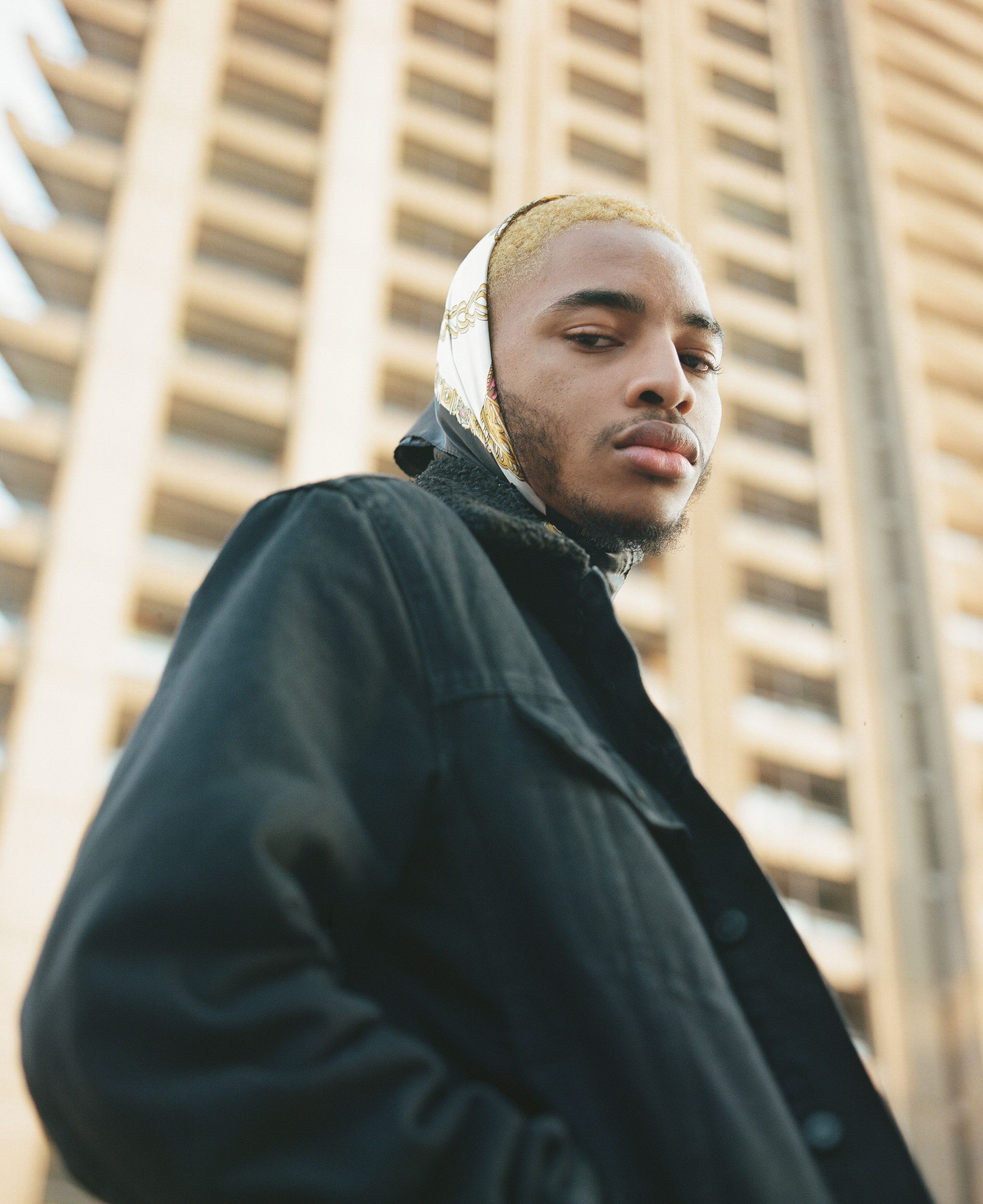 A man in a black jacket and gold and black headscarf stands in front of a modern building.