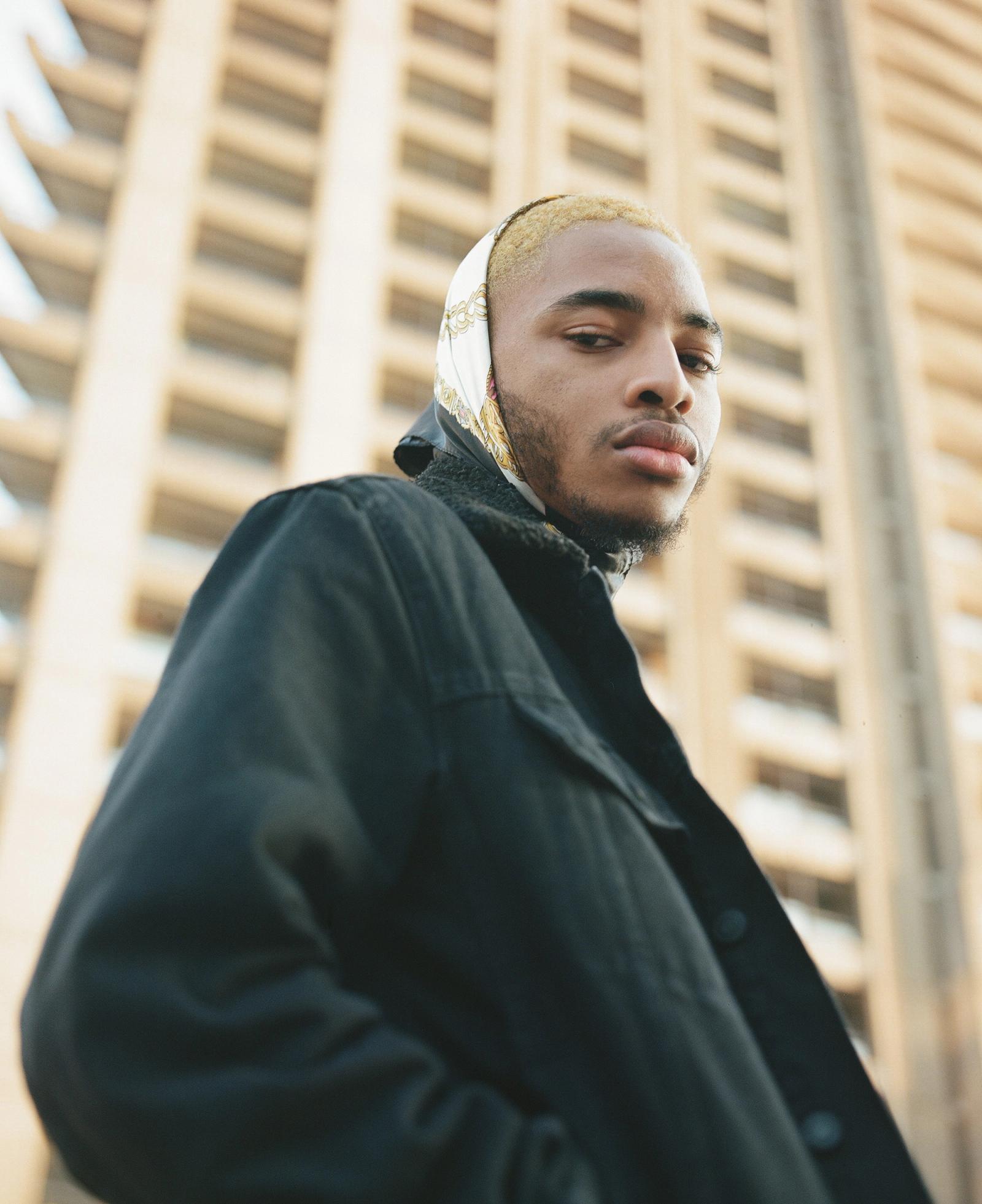 A man in a black jacket and gold and black headscarf stands in front of a modern building.
