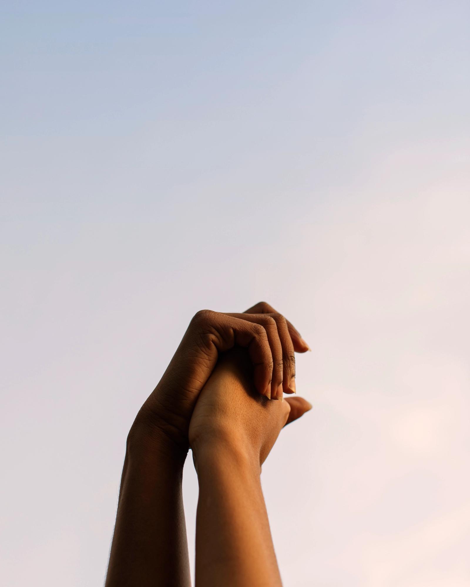 Two dark-skinned hands clasped together in a gesture of unity or prayer, with fingers intertwined and palms facing upwards.