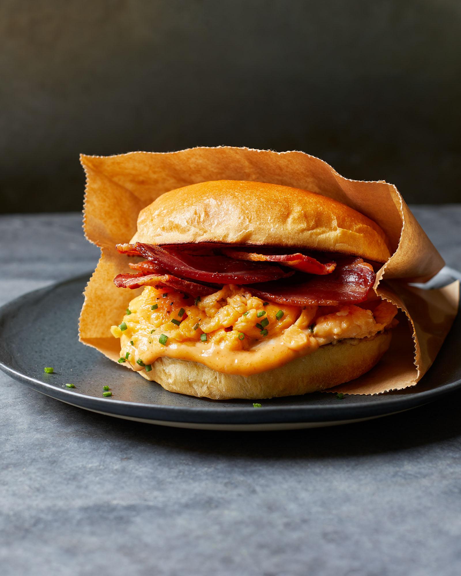 A toasted, open-faced breakfast sandwich with scrambled eggs, crispy bacon, and melted cheese on a toasted, flaky, golden-brown croissant, served on a dark grey plate.