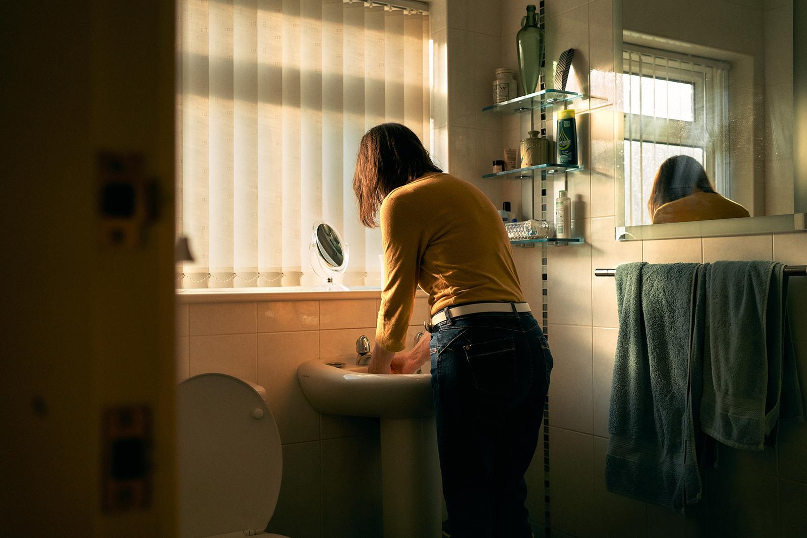 A woman in a yellow shirt and jeans is standing in a bathroom, washing her hands under a sink.
