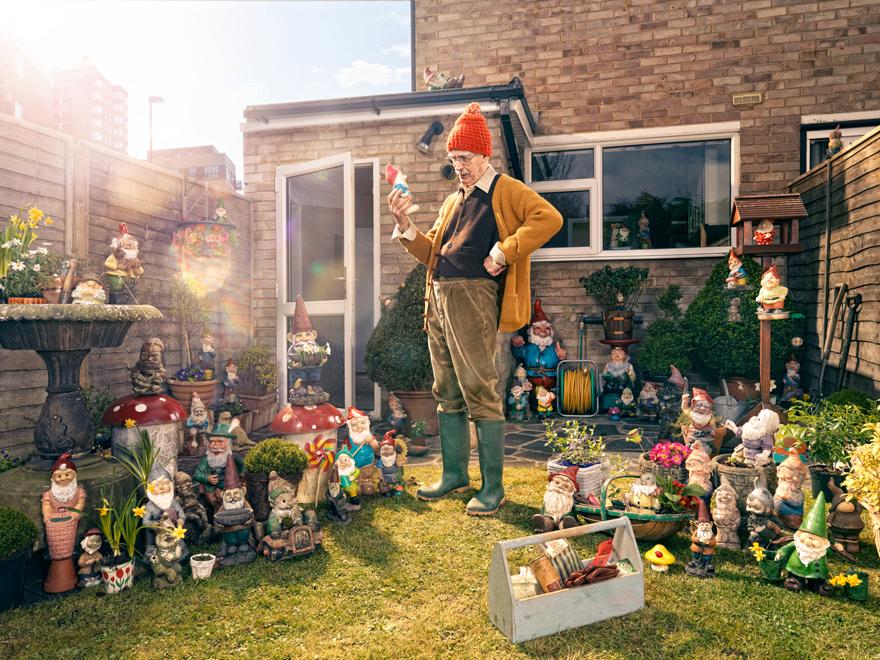 A man in a yellow cardigan, orange hat, and green wellies stands in a garden surrounded by numerous garden gnomes, some of which are holding flowers or other objects.