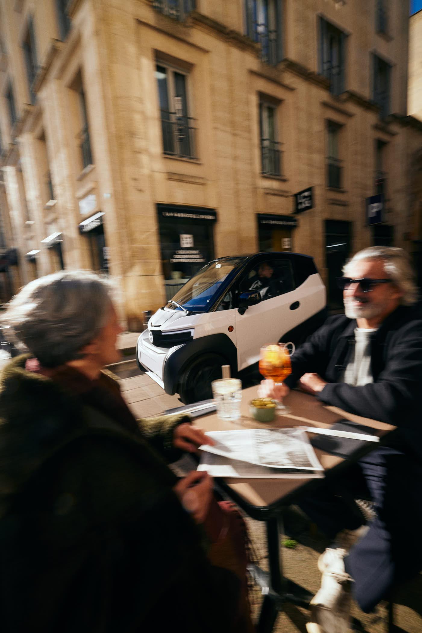 An elderly couple sits at a small outdoor table in front of a historic building. The man is holding a glass of orange drink, while the woman looks at a menu. A white car is.