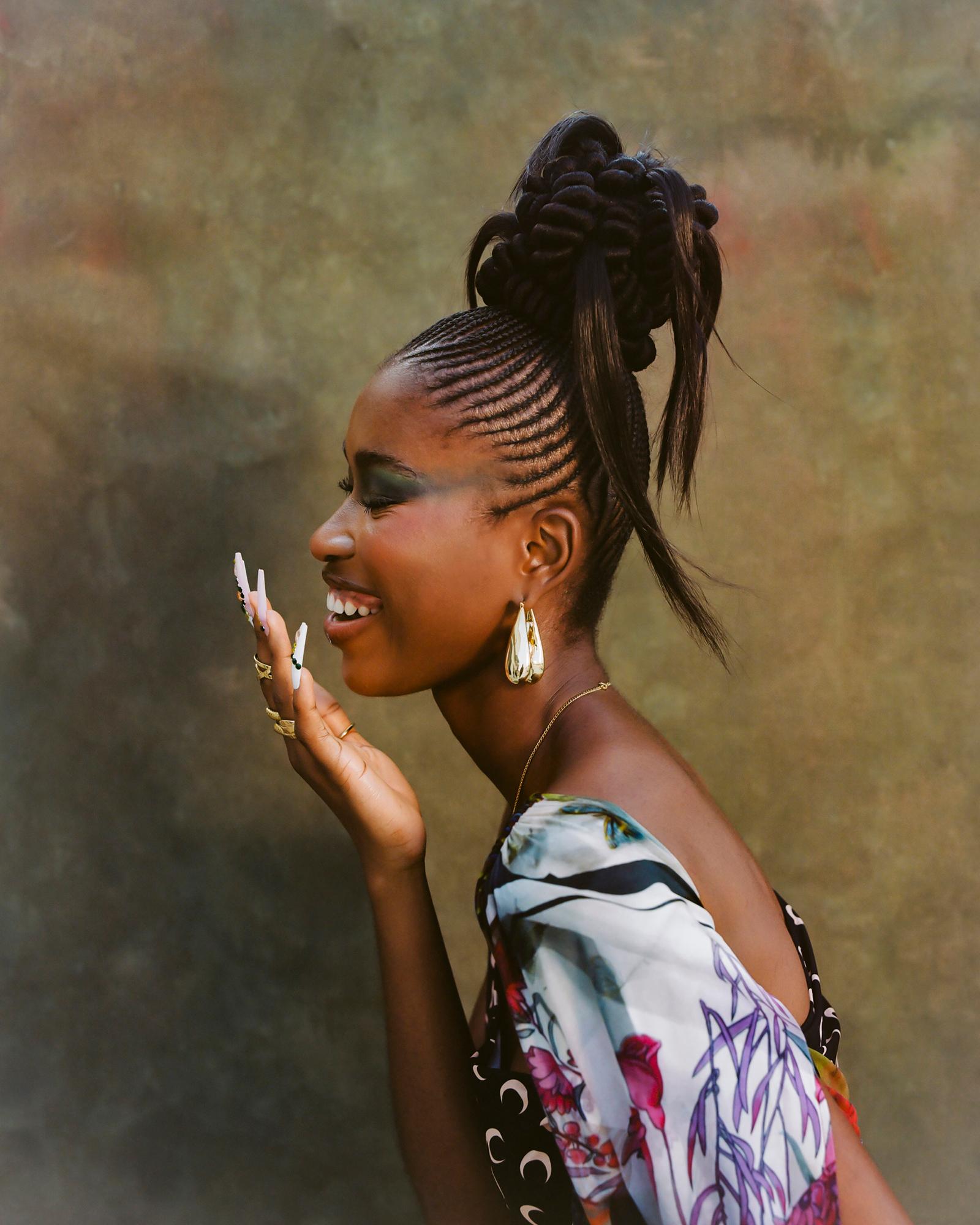 A woman with a high ponytail and braided cornrows, wearing a white and black floral top, smiling and looking to the side.