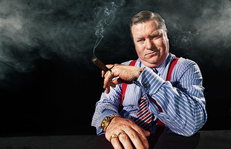 A man in a blue and white striped shirt, red suspenders, and a striped tie, holding a cigar and smiling at the camera.