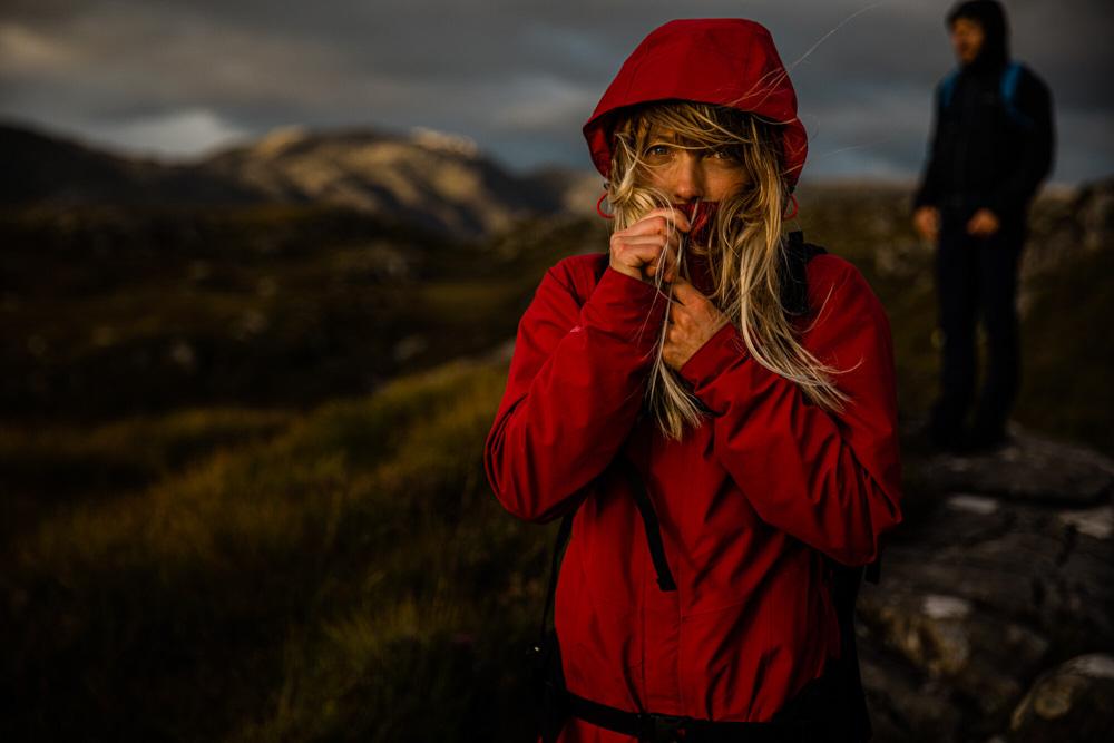 A woman in a red jacket and backpack stands on a rocky outcrop, gazing out at a mountainous landscape.