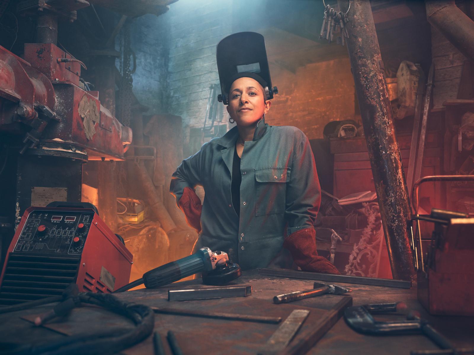 A woman wearing a welding helmet and protective gear stands in a dimly lit workshop surrounded by welding equipment.
