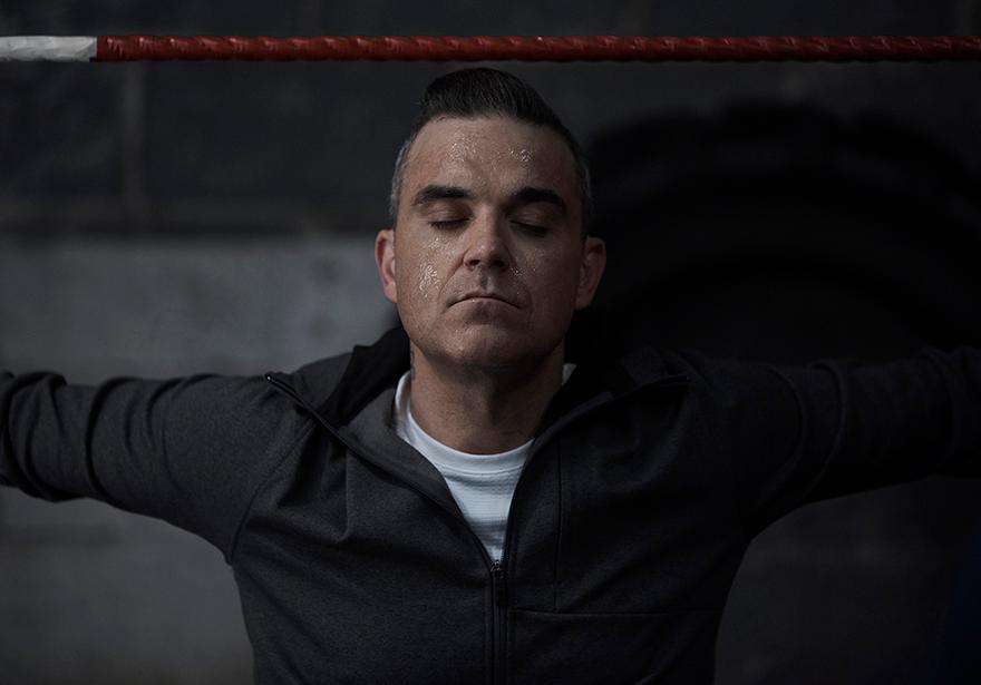 A man in a dark grey zip-up hoodie with a white t-shirt underneath, standing in front of a red and white rope, possibly in a gym or sports facility.