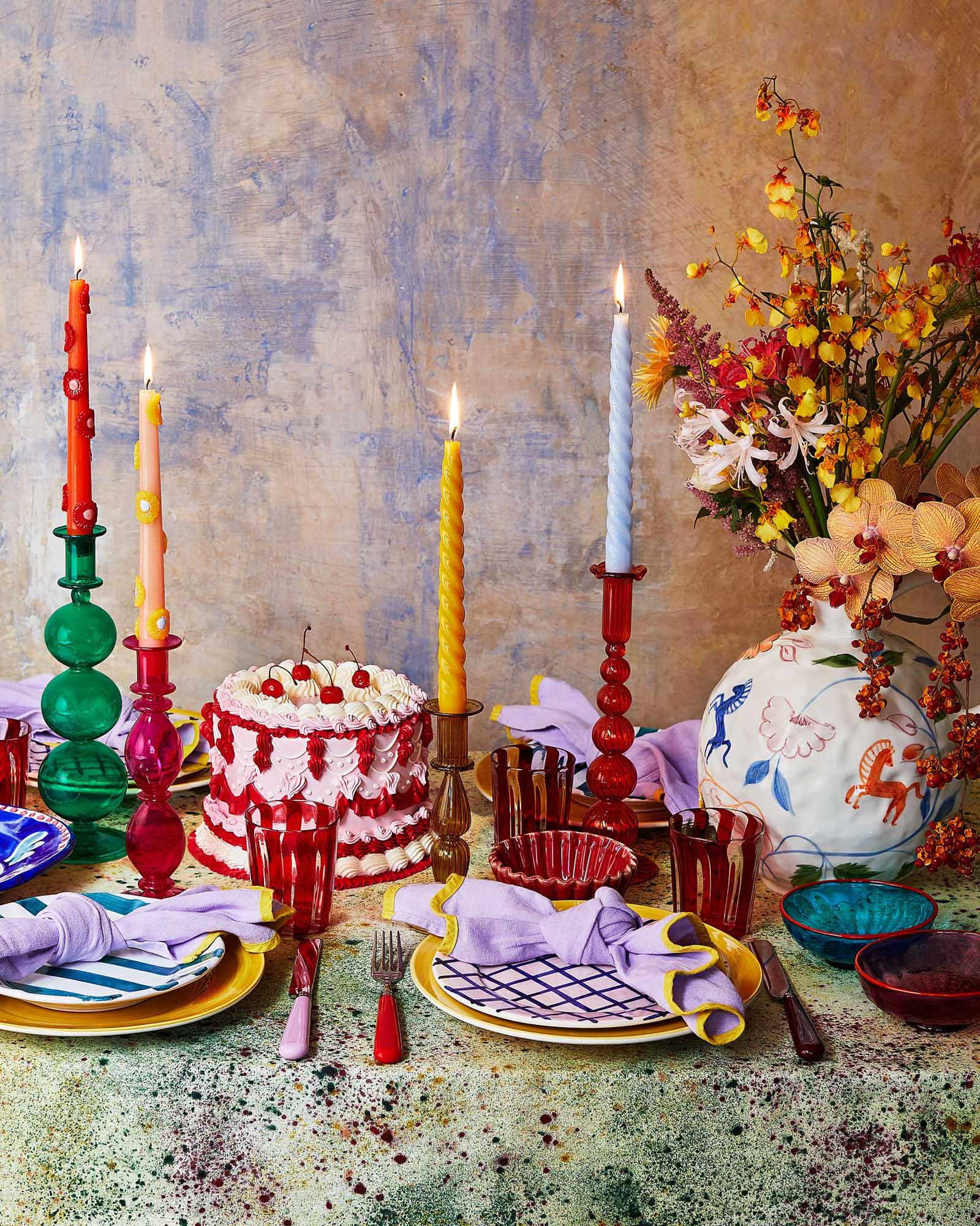 A table setting with a cake, flowers, and various tableware, including plates, glasses, and candles, set against a textured, abstract background.