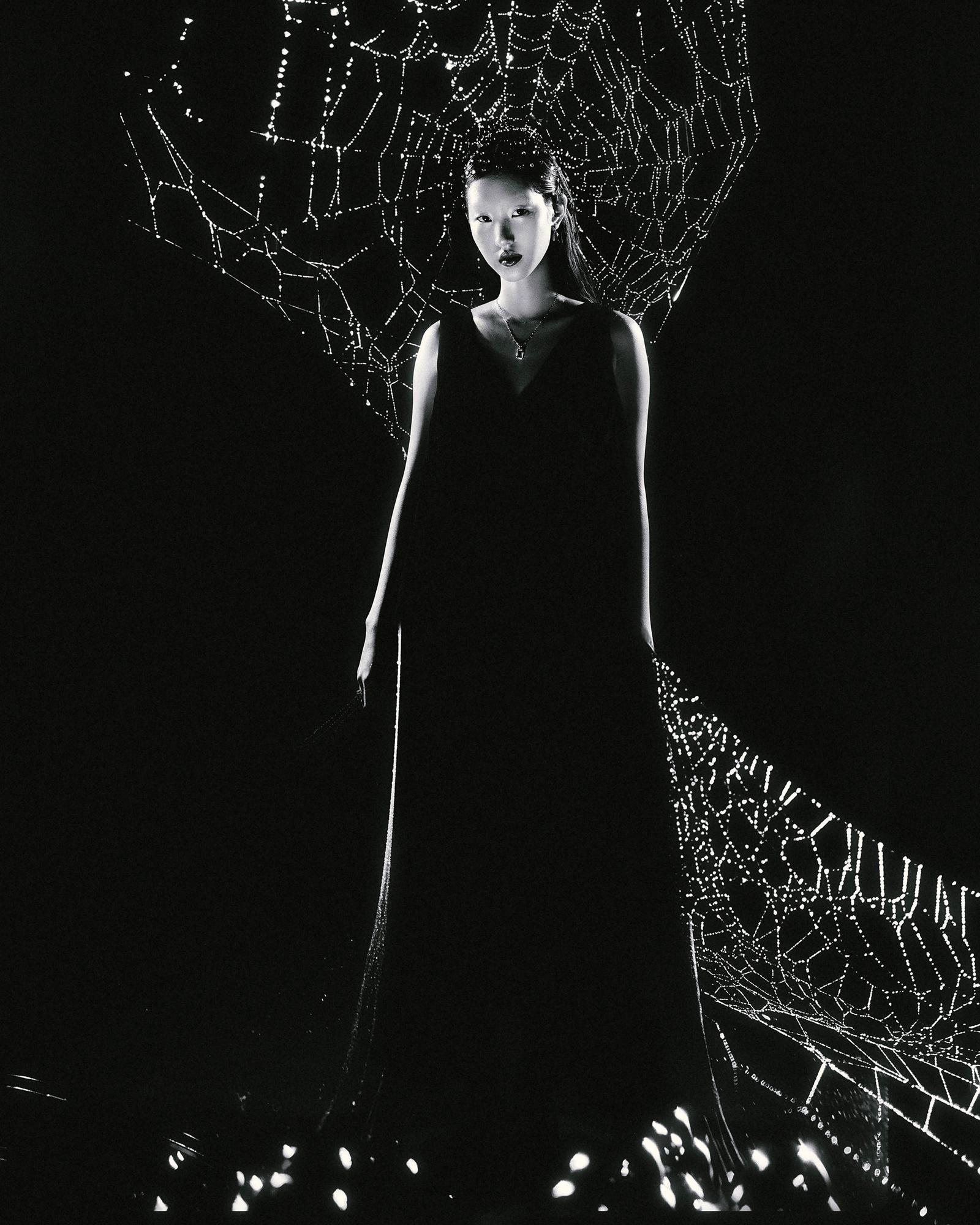 A woman in a dark, flowing dress stands in front of a massive spider web, illuminated by a faint, eerie glow.
