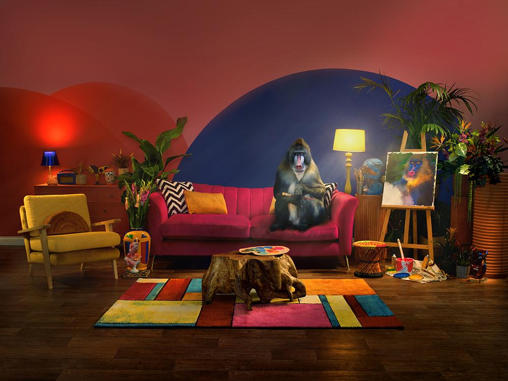 A monkey sits on a red couch and paints a woman's portrait surrounded by art supplies.
