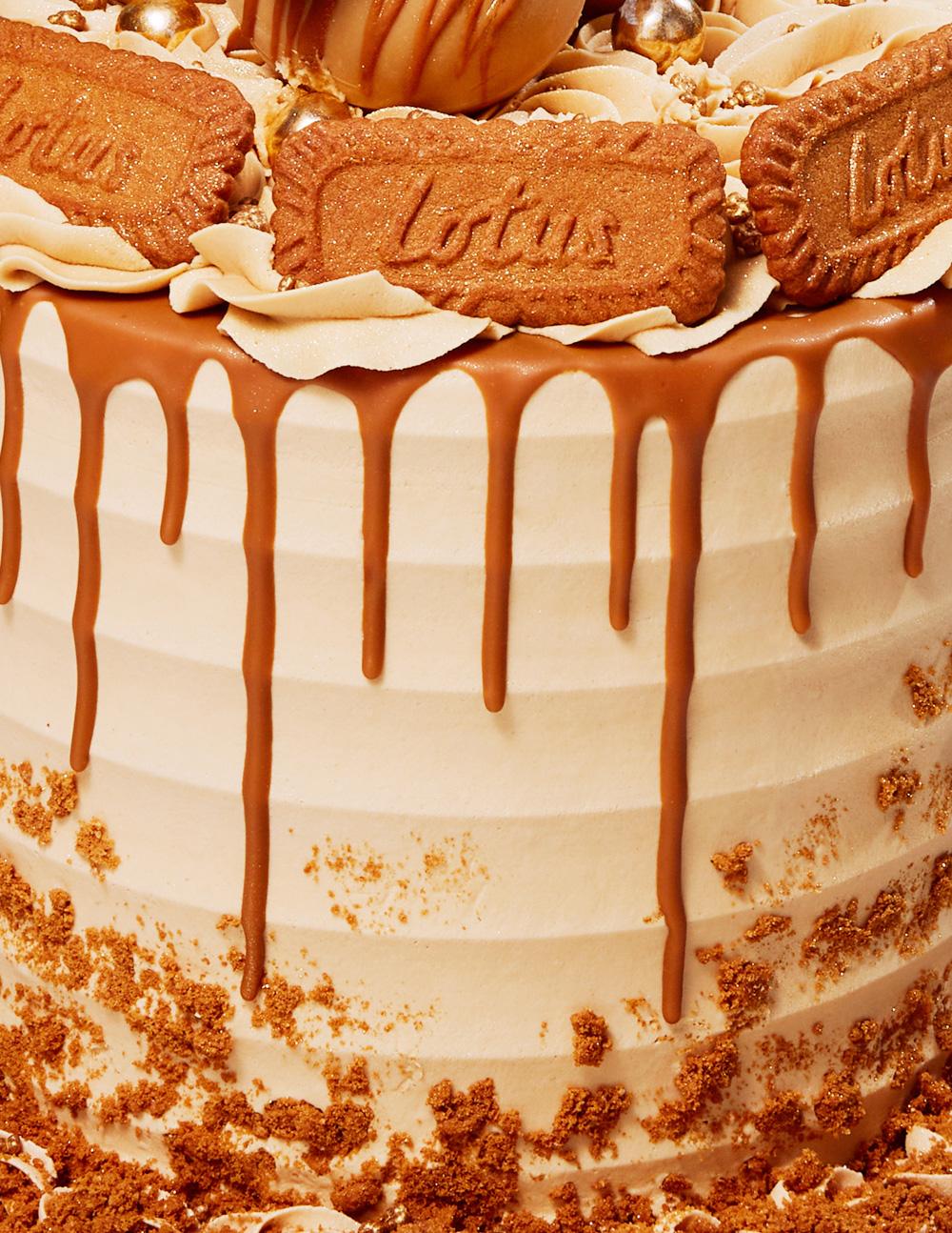 A close-up of a large, multi-layered cake with a light brown, creamy frosting, topped with Biscoff biscuits and a chocolate drizzle.