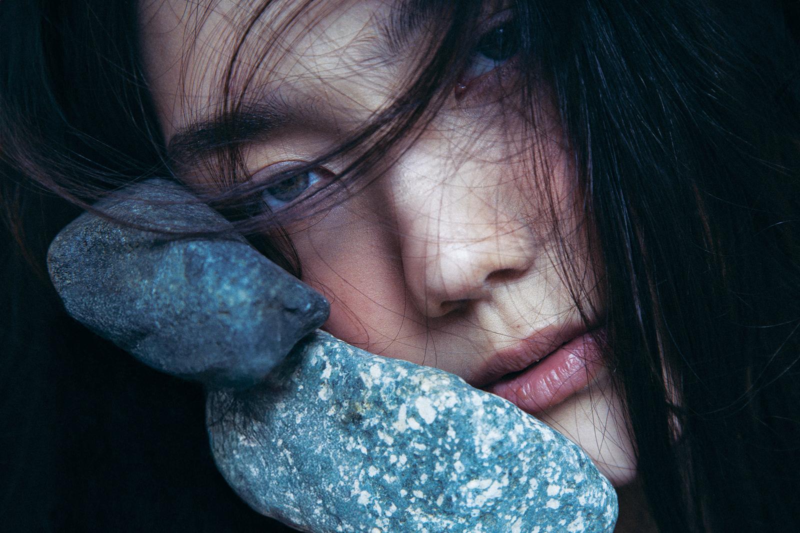 A woman with long, dark hair and a pale complexion, holding a large, rough stone in her hand, with a subtle, introspective expression.