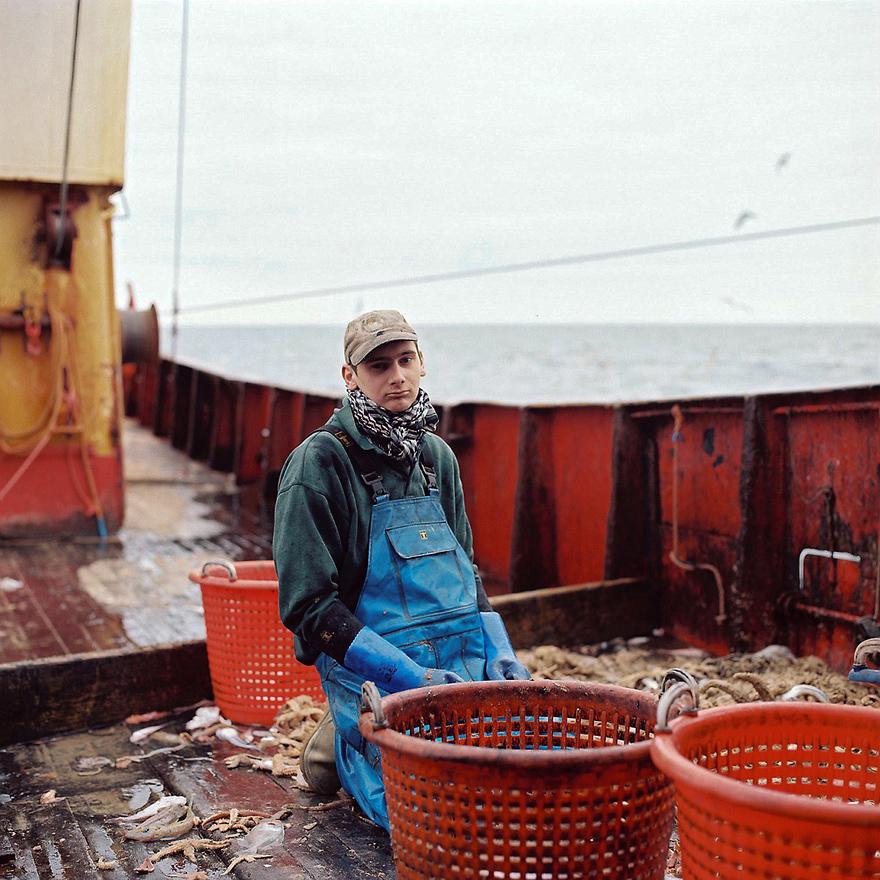 A man in a green sweatshirt, blue overalls, and a scarf is kneeling on a boat, surrounded by fishing nets and baskets, with a large red container in the background.