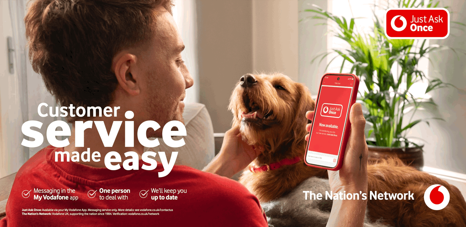 A man in a red shirt is sitting on a couch with a dog, holding a smartphone with the Vodafone app on the screen.