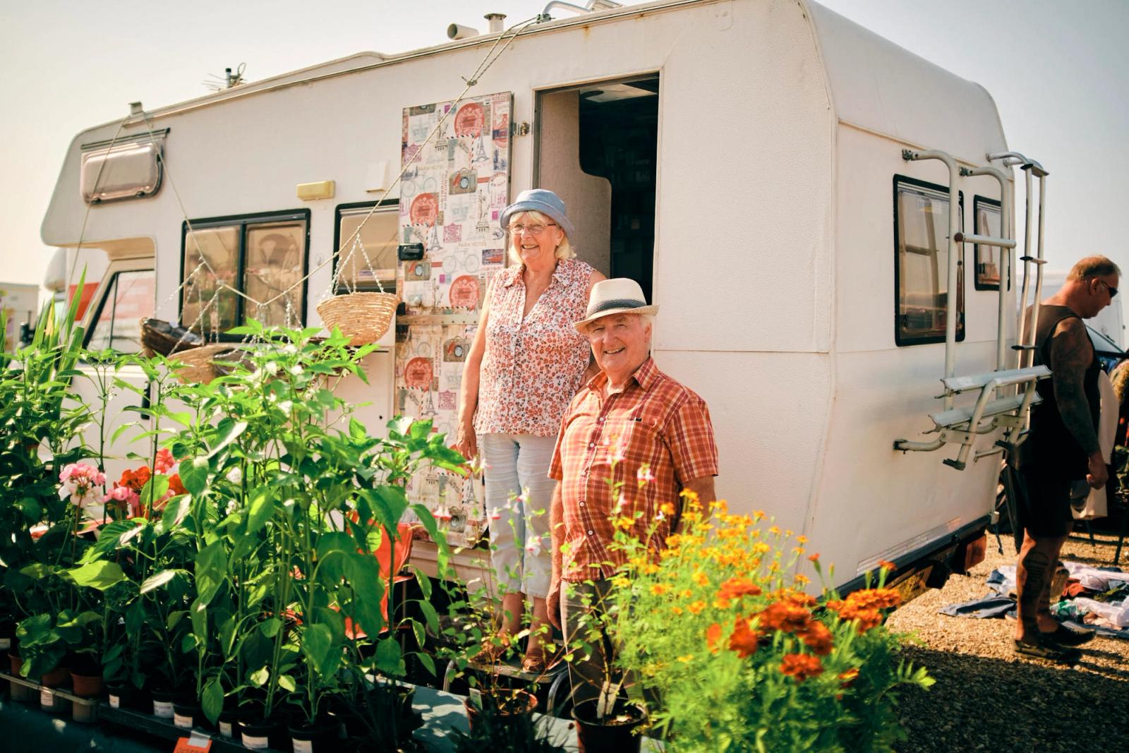An older couple standing in front of a camper van, surrounded by potted plants and flowers. They are both smiling and looking directly at the camera.