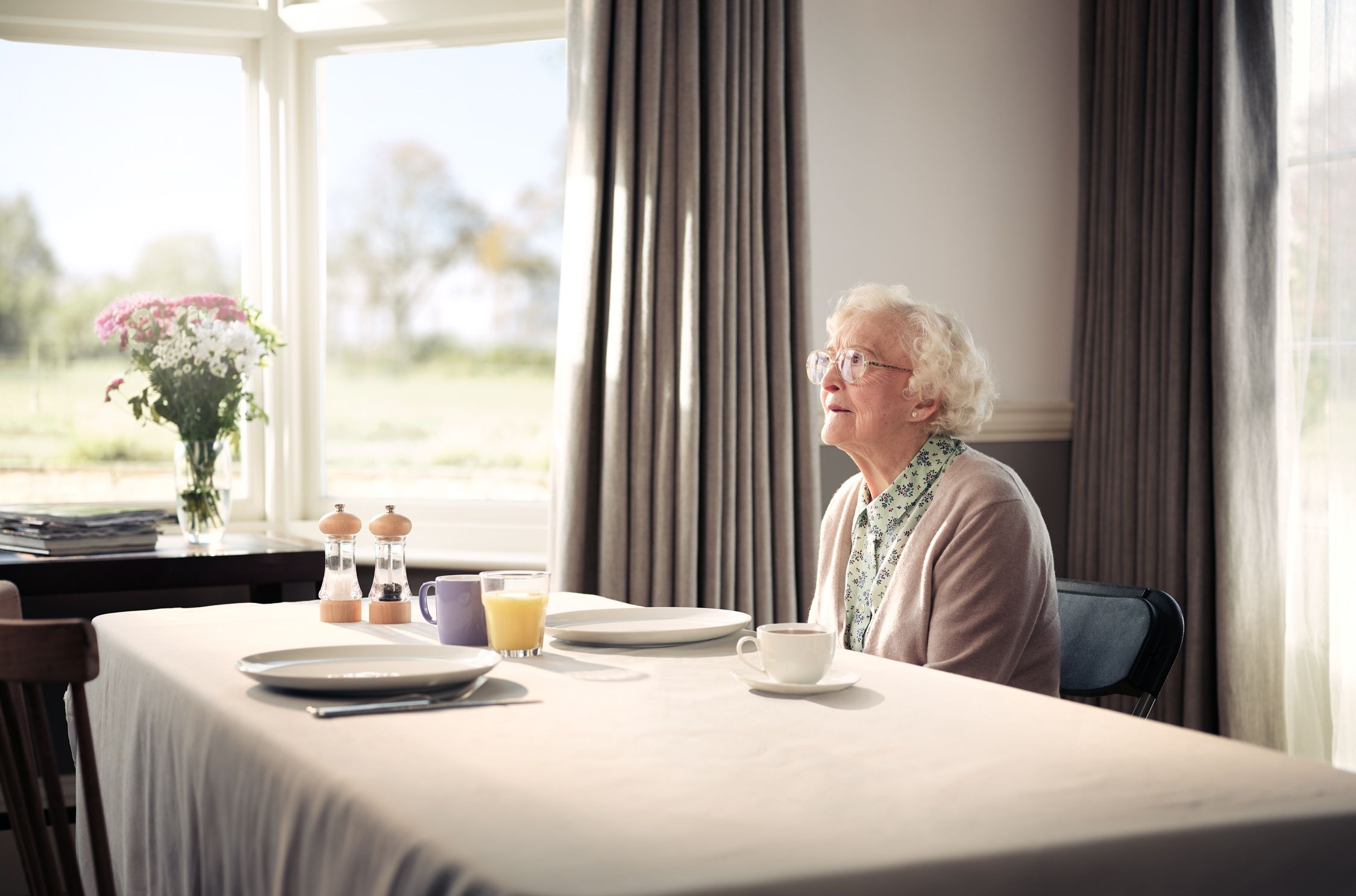 An elderly woman sits at a table in a well-lit room, looking out the window at a garden or park in the distance, with a table setting in front of her.