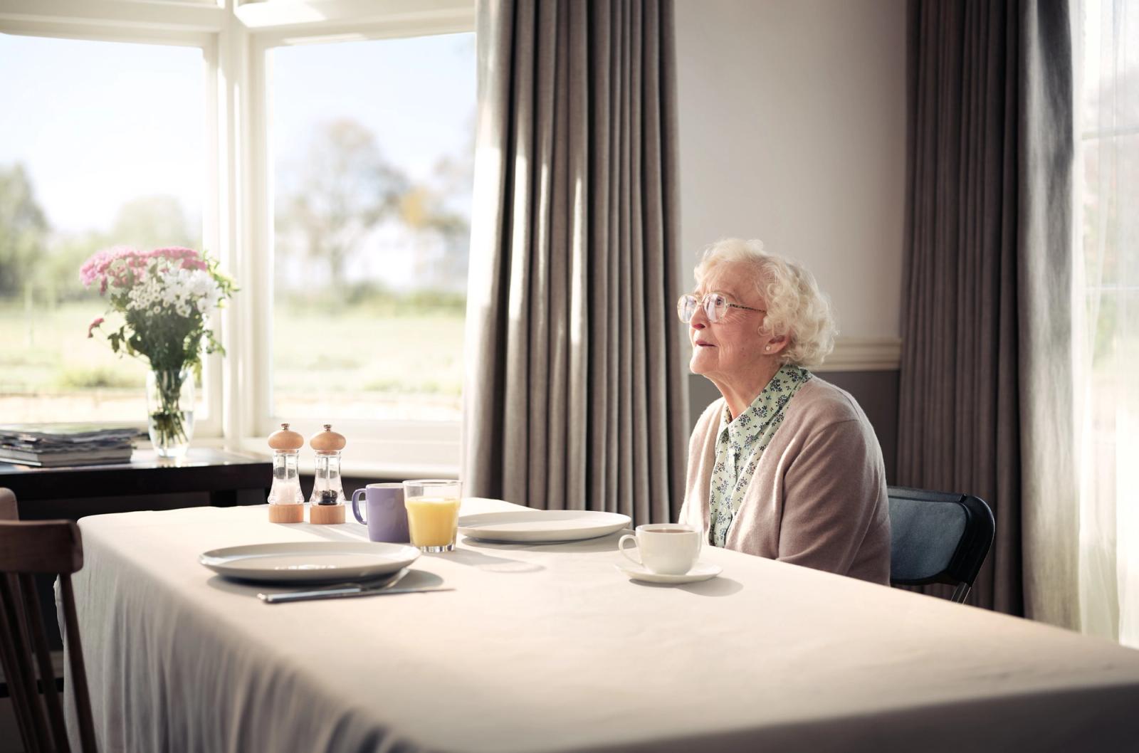 An elderly woman sits at a table in a well-lit room, looking out the window at a garden or park in the distance, with a table setting in front of her.