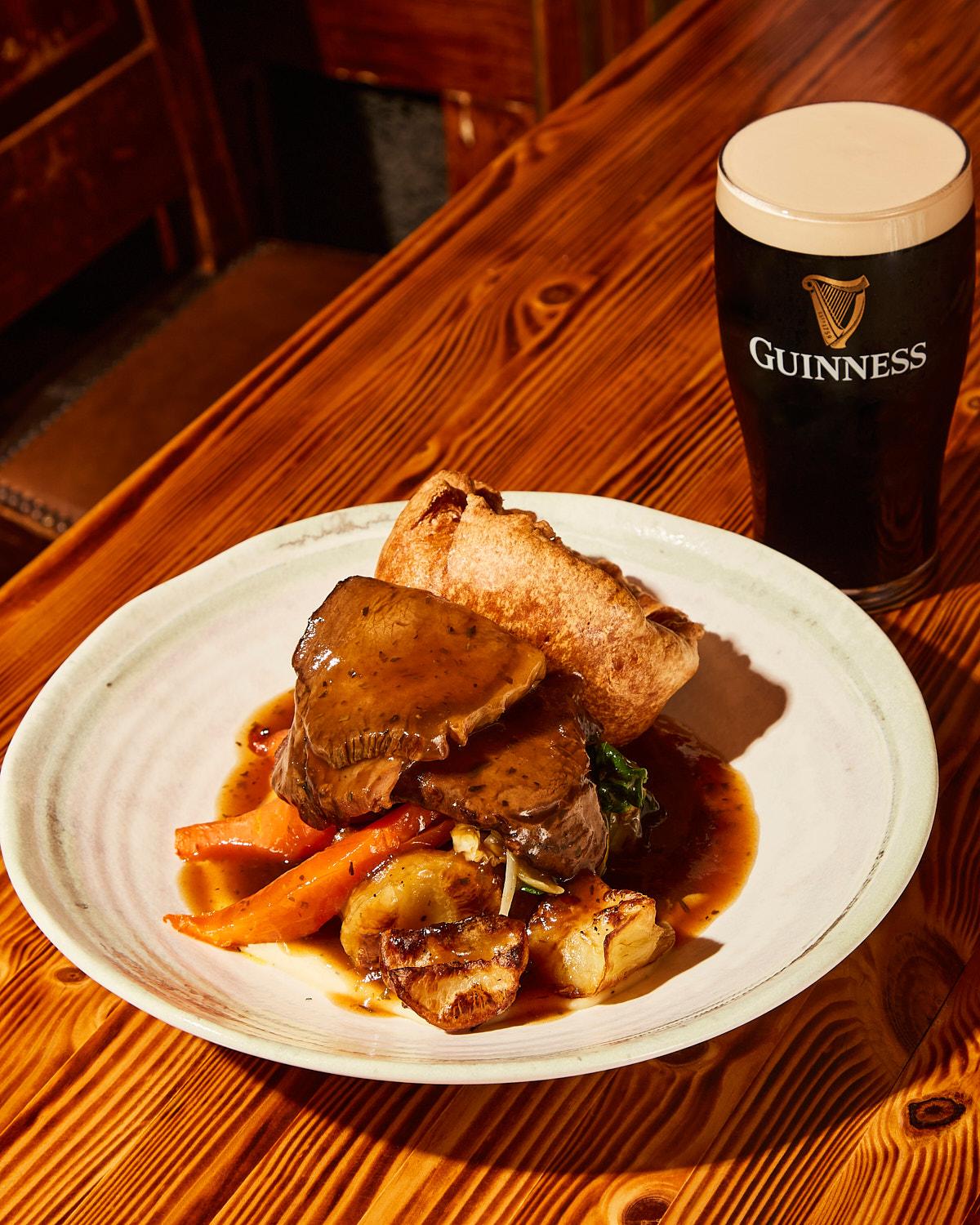 A plate of food, including a beef dish, vegetables, and a pastry, served with a glass of Guinness beer on a wooden table.