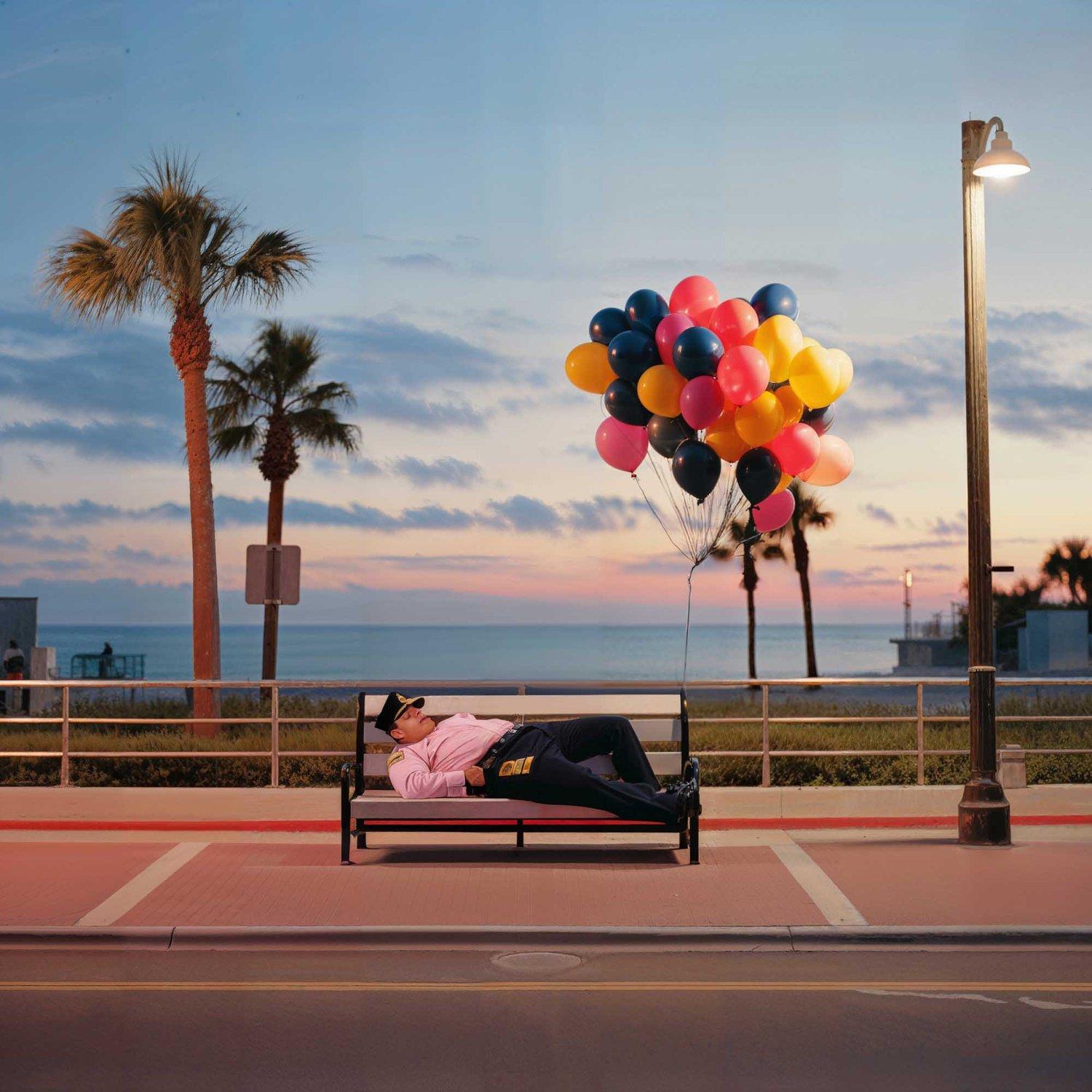 A police officer is sleeping on a bench, surrounded by balloons and a beach in the background.