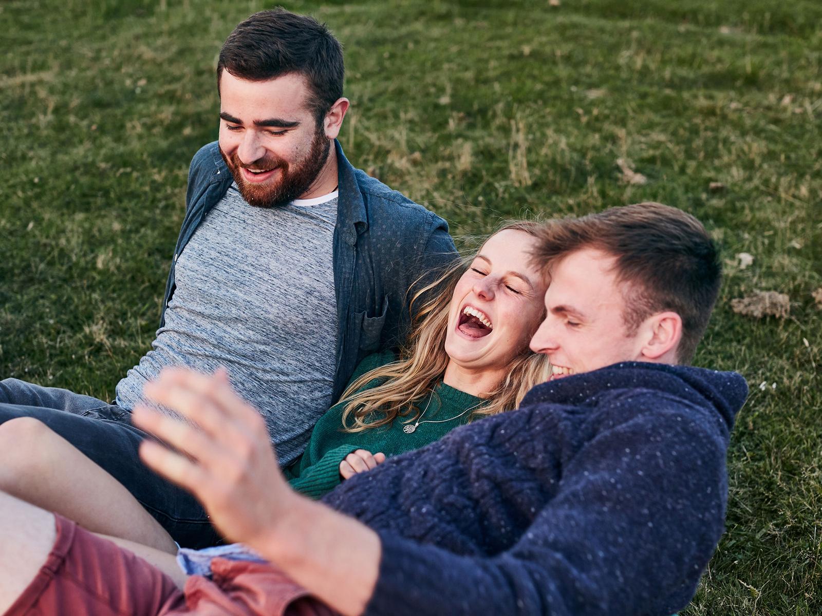 Three people laughing and hugging in a green field, each wearing different colored shirts.