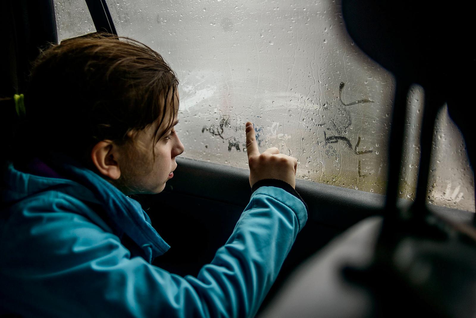 A young girl with wet hair and a blue jacket is drawing on a rainy bus window.