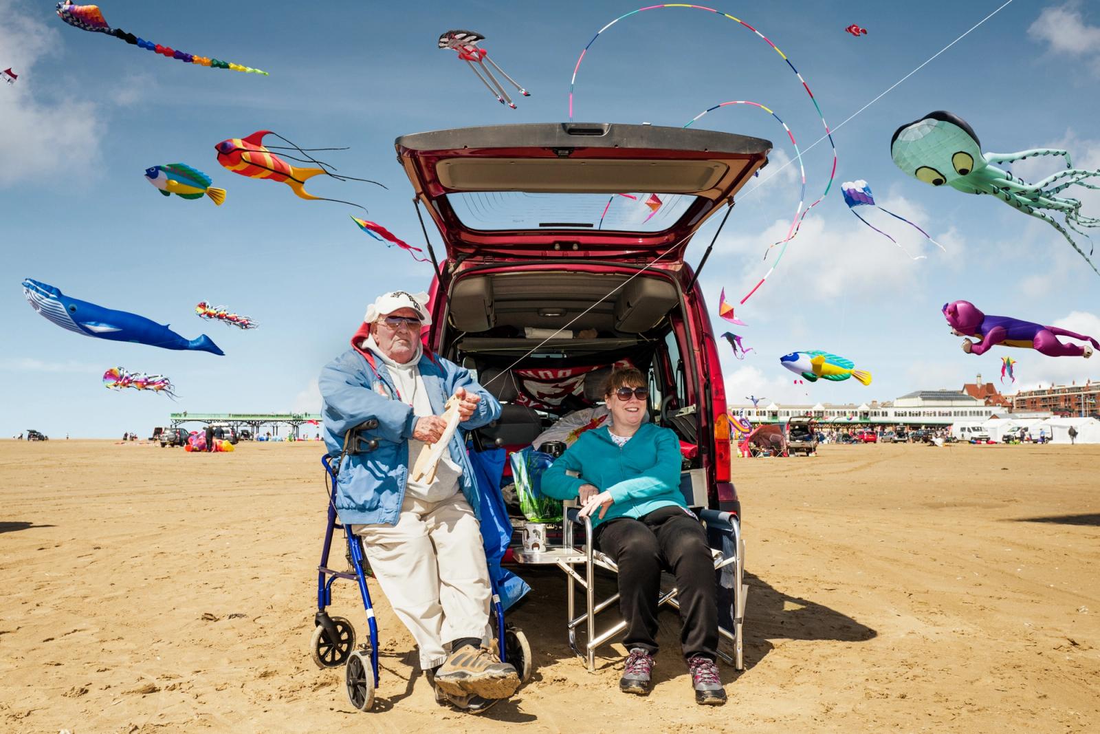 An older man in a blue jacket and white pants is sitting in a wheelchair on a beach, surrounded by kites and a red car.