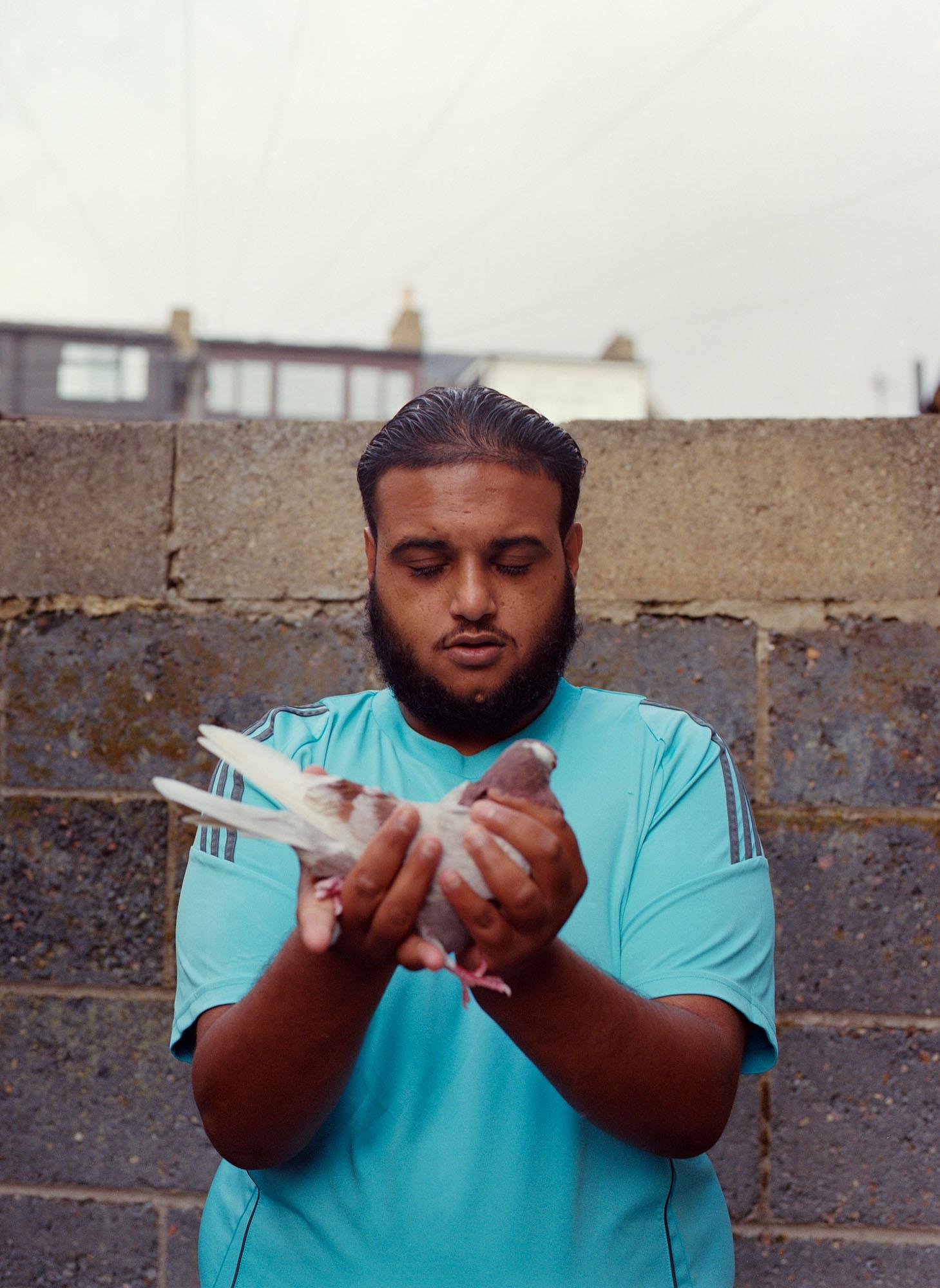 A man holding a white pigeon in front of a grey concrete wall, wearing a blue t-shirt.