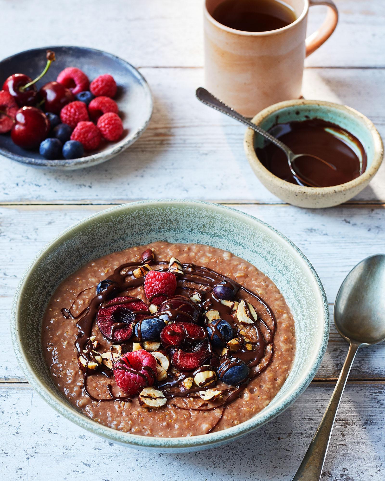 A bowl of chocolate oatmeal with fruit and nuts, next to a cup of coffee and a plate of berries.