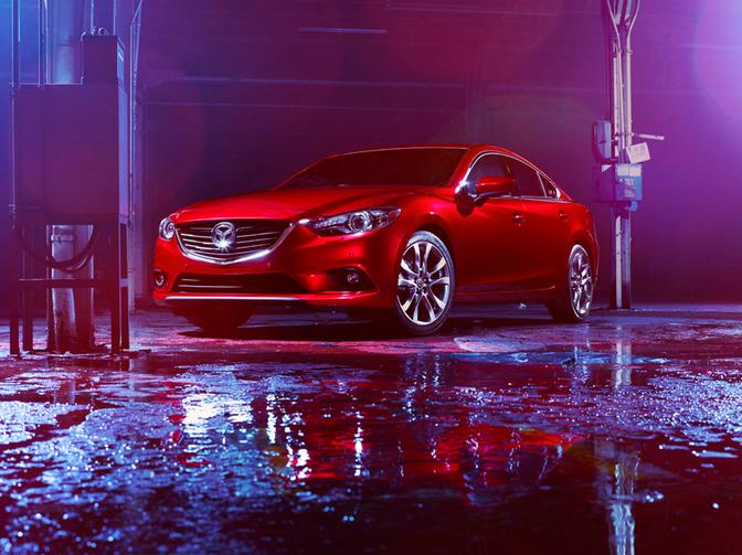 A red Mazda car is parked in a dimly lit, wet, and dark garage or car wash facility, with water and oil on the ground.