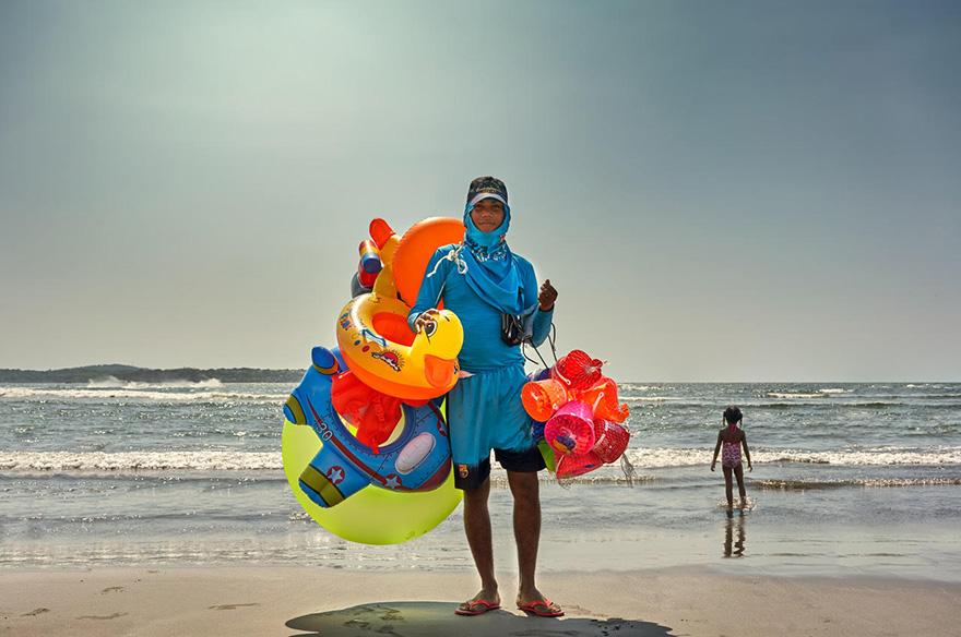 A man in a blue wetsuit and cap, holding a collection of brightly colored, inflatable beach toys, on a sunny beach with a child in the background.