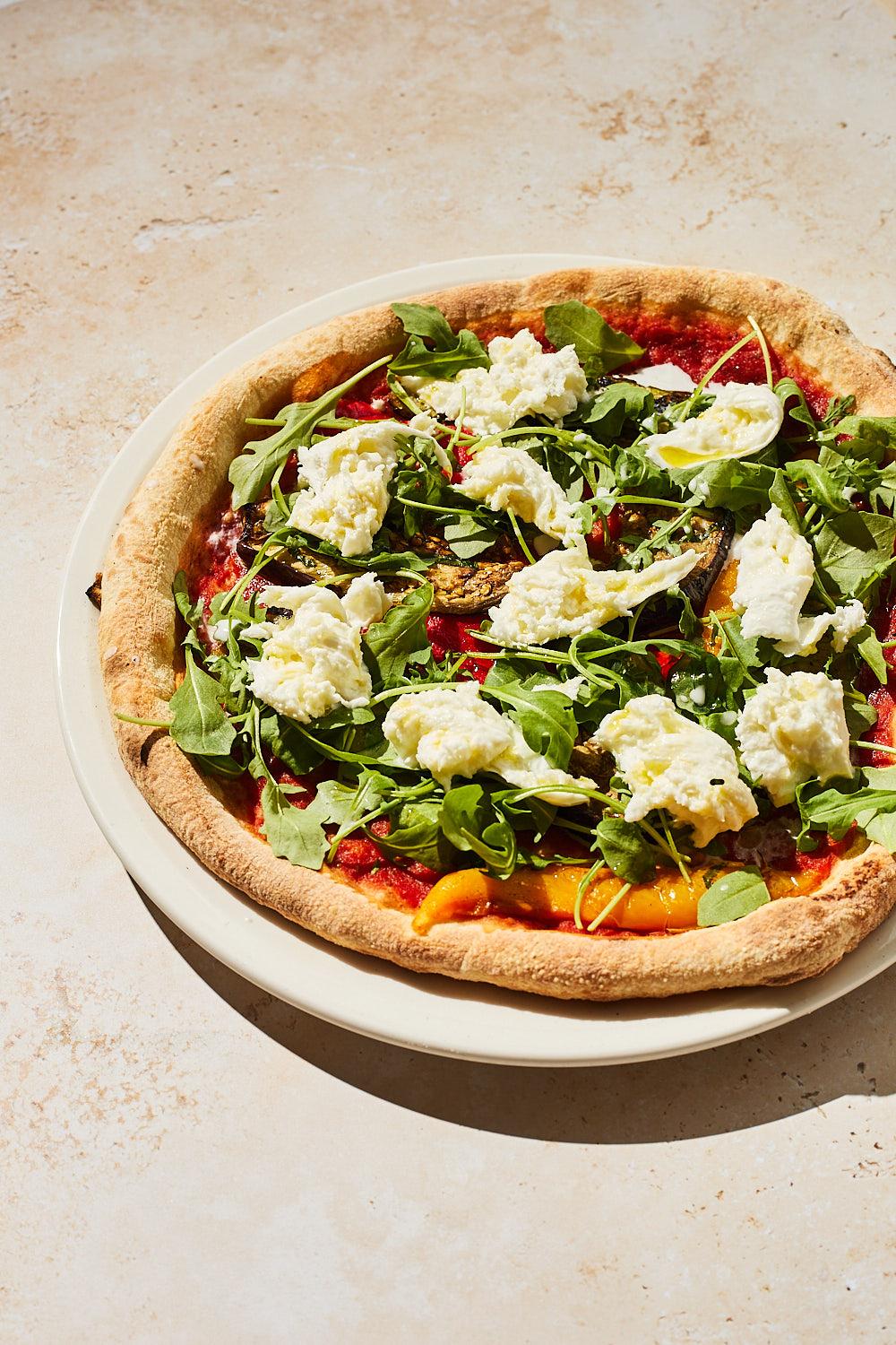 A round, flat, crispy pizza topped with various ingredients, including vegetables, cheese, and herbs, on a white plate.