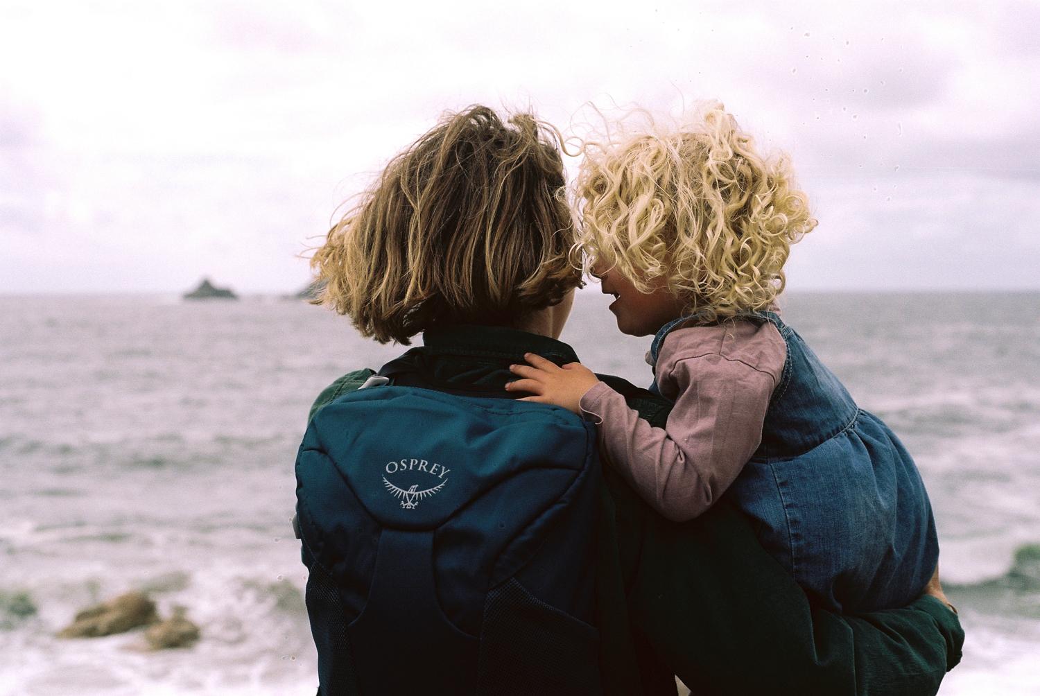 A woman in a blue jacket is holding a young child with curly blonde hair, both facing the ocean.