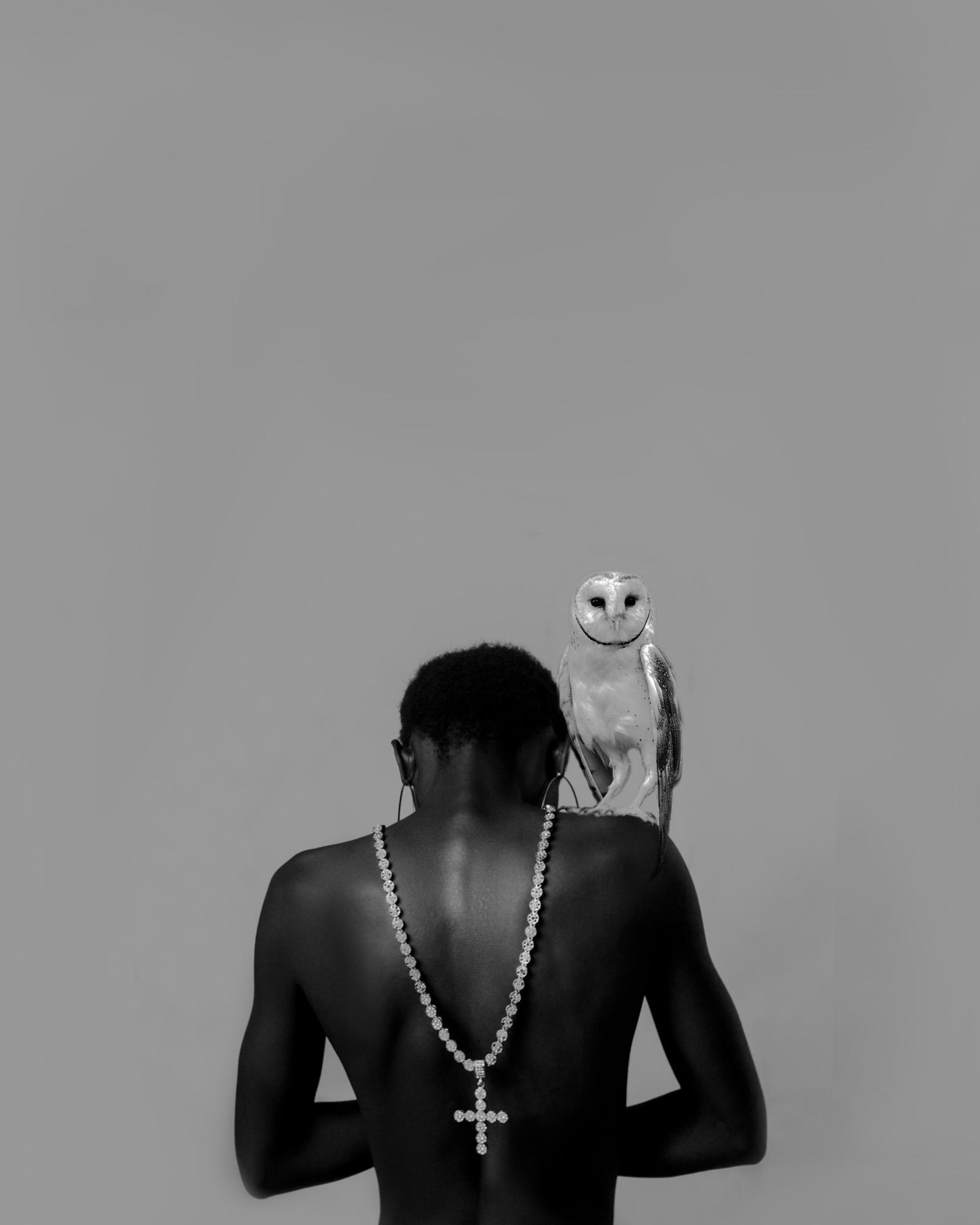 A person wearing a necklace with a pendant in the shape of an owl, standing in front of a plain background.