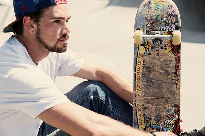 A man in a white t-shirt and jeans is sitting on the ground, holding a skateboard.