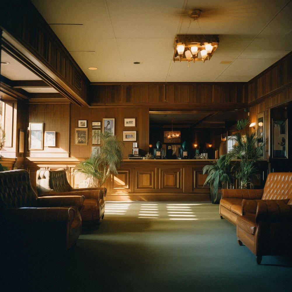 A dimly lit, empty lobby with a few chairs and a reception desk, with a large window on the left wall showing a city skyline at night.