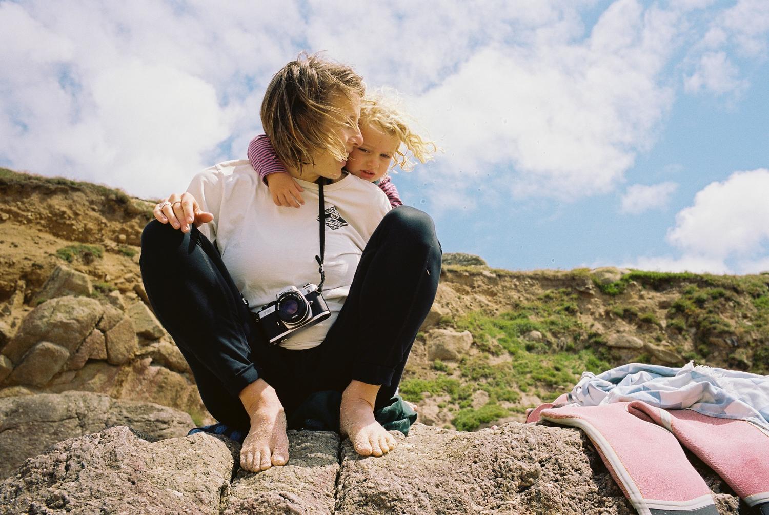 A woman, likely a mother, is sitting on a rocky outcrop, holding a young child, who is embracing her neck, in a scenic, outdoor setting, with a camera around her neck.