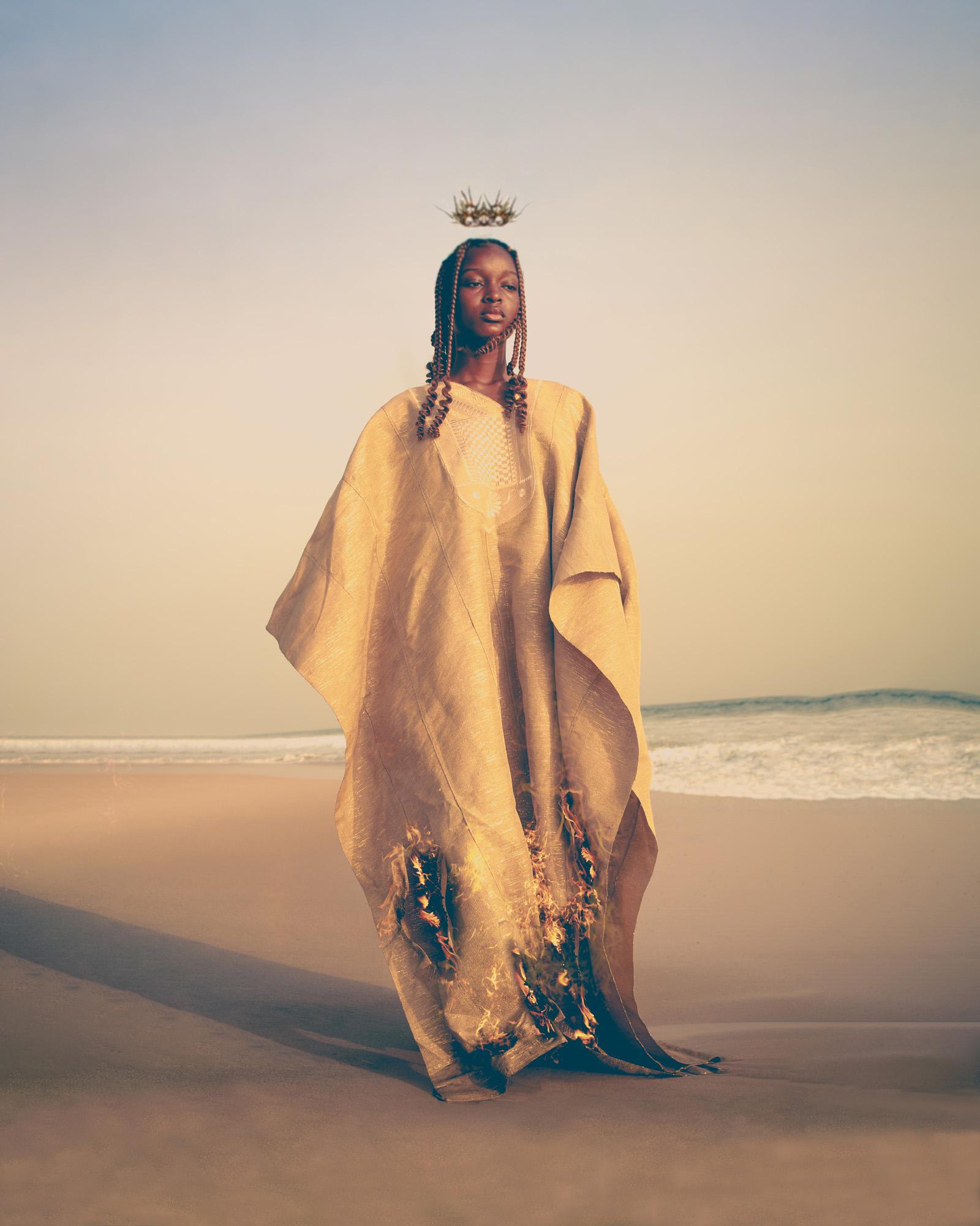 A woman wearing a traditional African garment stands on a beach with the ocean visible.