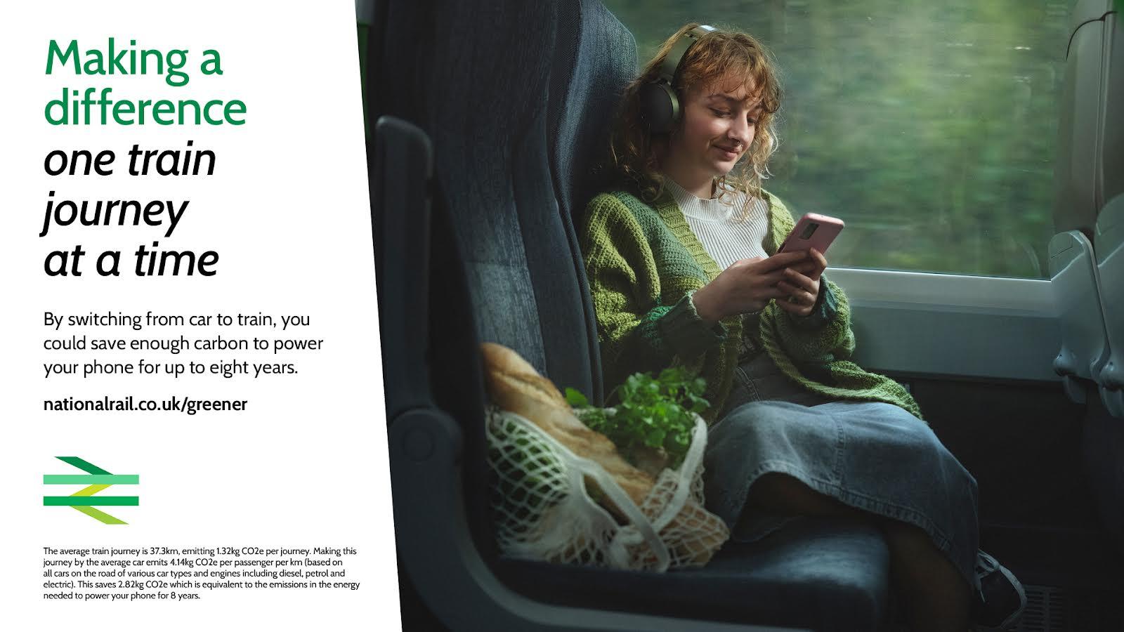 A woman sits on a train, wearing a green sweater, jeans, and headphones, looking at her phone.