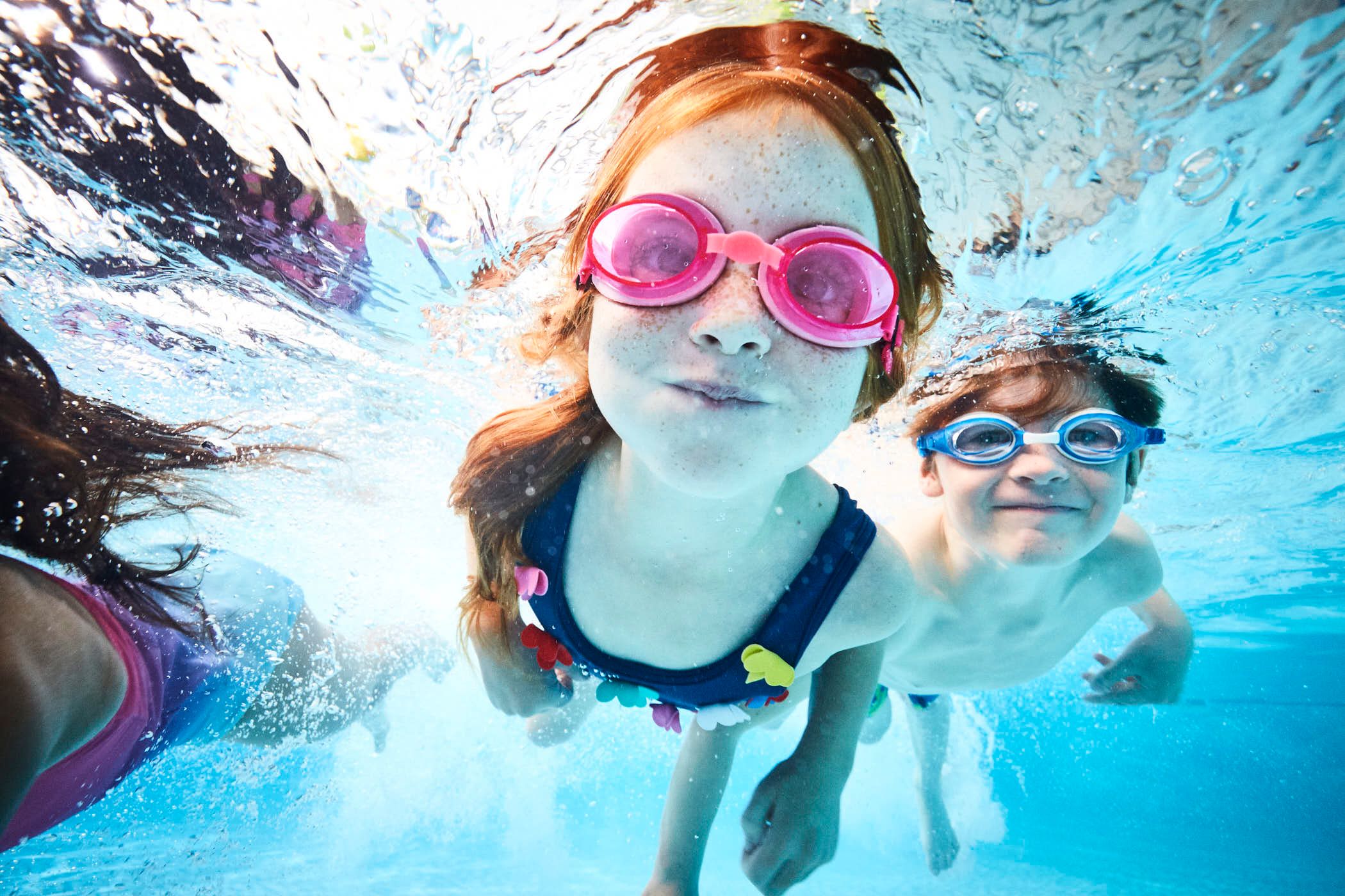 A young girl and boy are swimming underwater, wearing pink and blue goggles respectively, with a playful and mischievous expression on their faces. The girl has long red hair and.