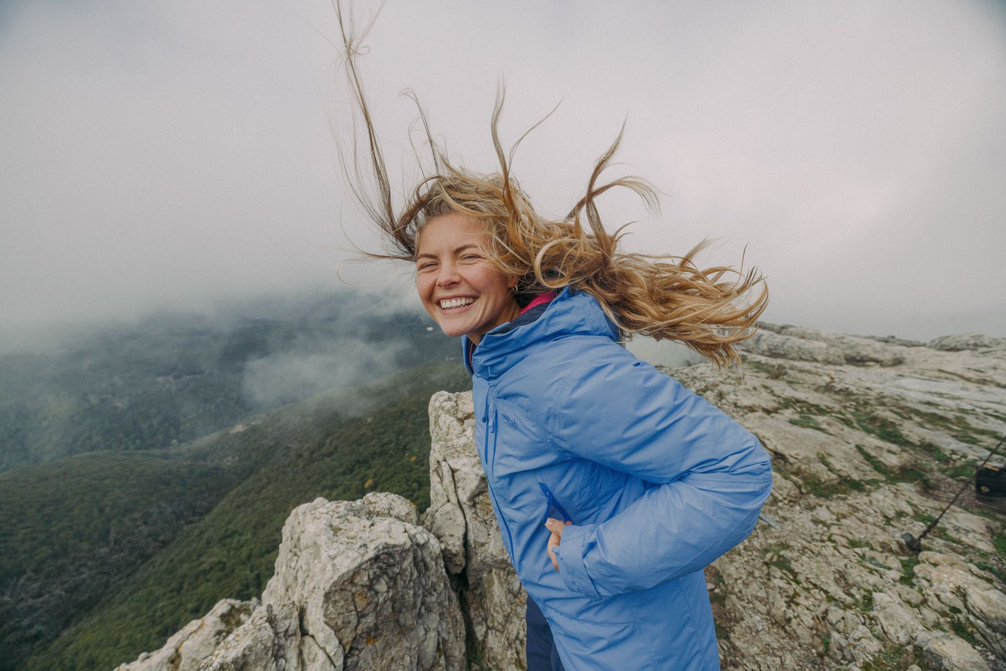 young woman with blonde hair blowing in the wind standing on top of the mountain with a blue jacket and smiling james bowden