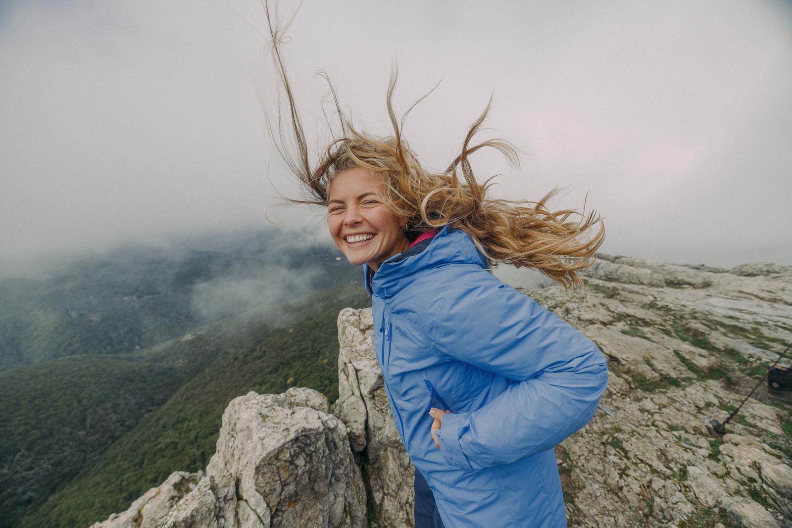 young woman with blonde hair blowing in the wind standing on top of the mountain with a blue jacket and smiling james bowden