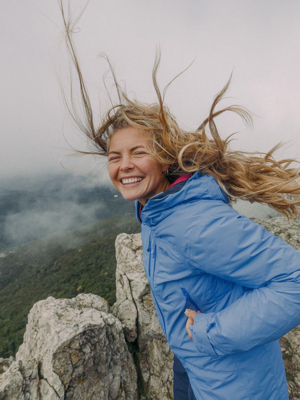 young woman with blonde hair blowing in the wind standing on top of the mountain with a blue jacket and smiling james bowden