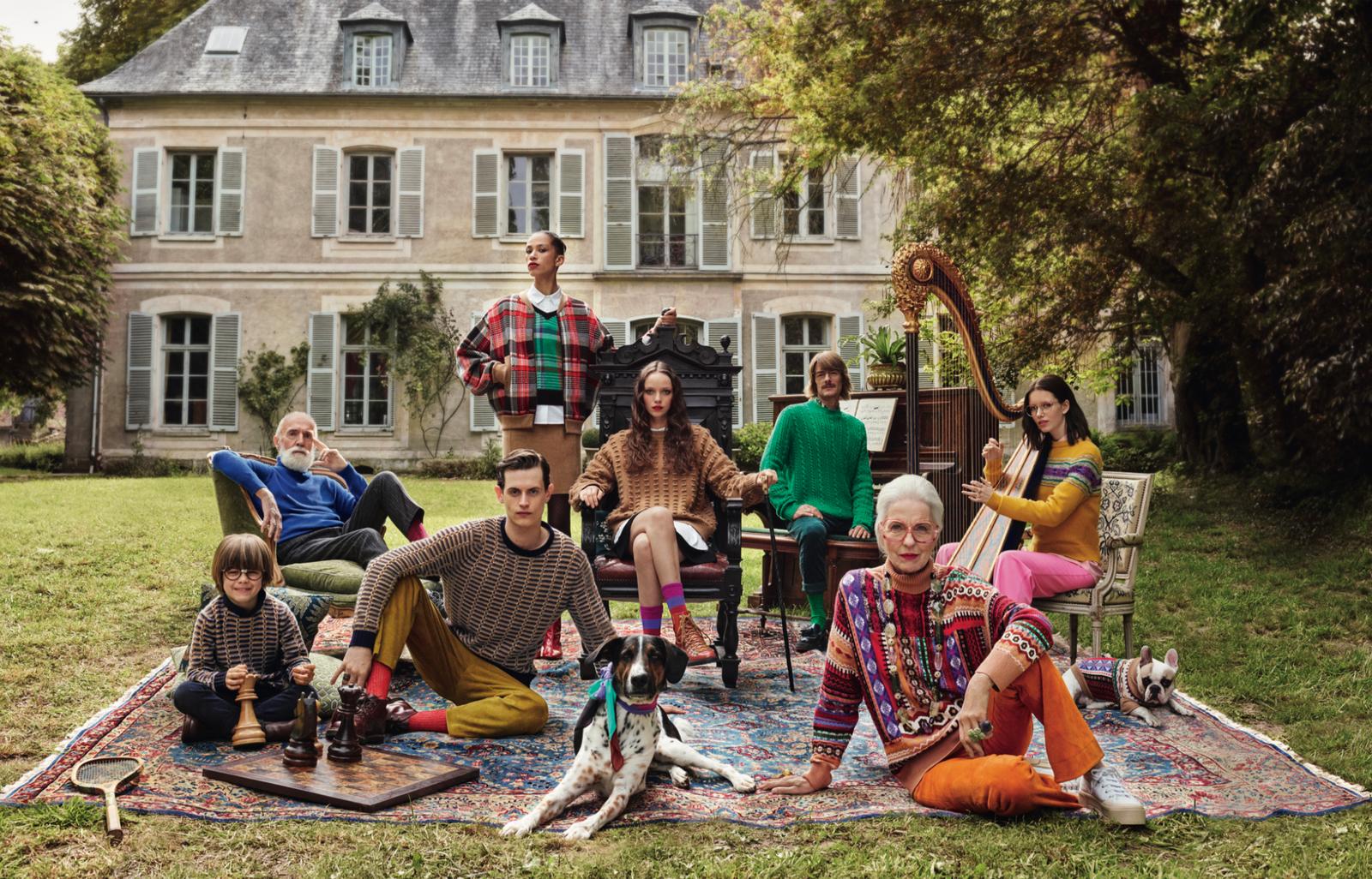 A group sits on a rug in front of an old house, dressed in mixed modern and vintage attire, with some playing a harp and others playing chess.