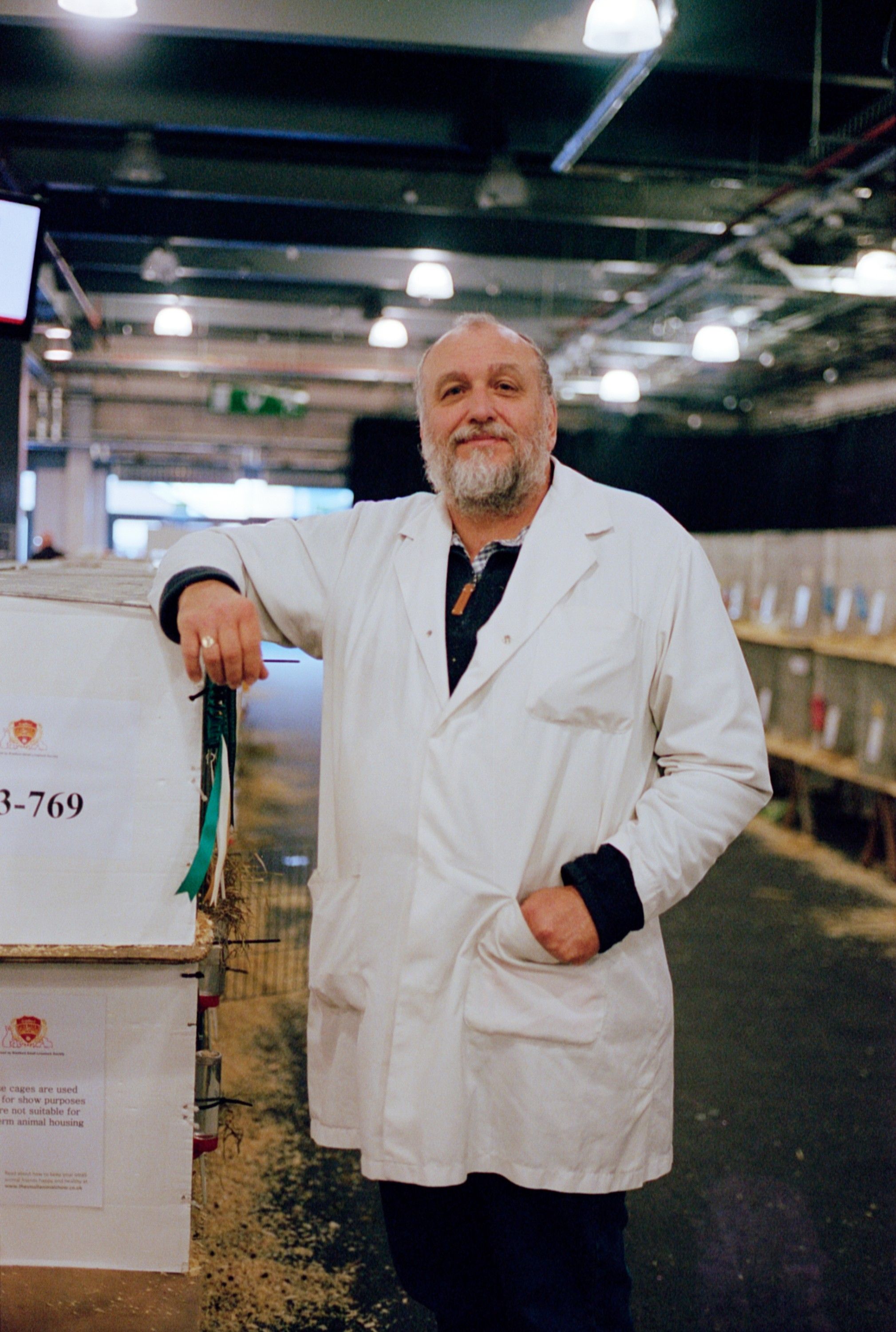 A man in a white lab coat stands in a large, industrial-style room with rows of animal cages in the background.