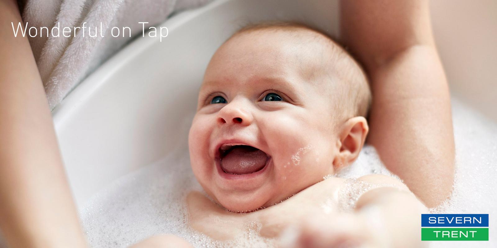 A baby in a bathtub, surrounded by soap bubbles and water, smiling and looking up at the camera.