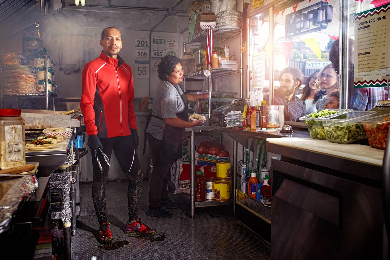 A man in a red jacket and black pants stands in a food truck kitchen, surrounded by food preparation equipment and a few customers in the background.