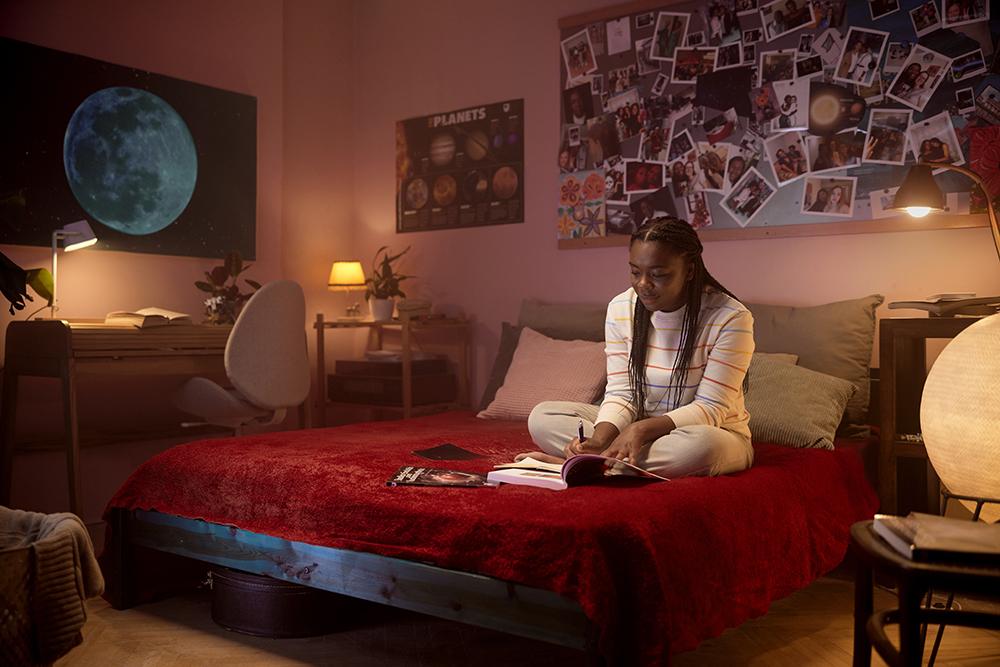 A young girl with long braids sits on a bed, surrounded by books and a laptop, in a room with a wall of posters and a desk with a computer and a chair.